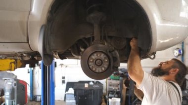 Üniformalı tamirci atölyeye yeni bir parça takıyor. İşçi otomobilinden kemer değişimine. Garajdaki kaldırma aracının altında profesyonel sakallı oto tamircisi çalışıyor. Yavaş çekim.