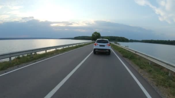 汽车在湖边的公路上行驶 夏天的一天 白色的Suv沿着堤坝的路线行驶 跟随汽车通过河桥 风景的方式 旅行或旅程的概念 空射慢速弹药 — 图库视频影像