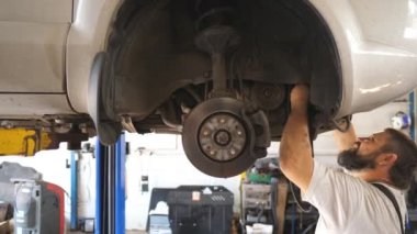 Üniformalı tamirci atölyeye yeni bir parça takıyor. İşçi otomobilinden kemer değişimine. Garajdaki kaldırma aracının altında profesyonel sakallı oto tamircisi çalışıyor. Yavaş çekim.