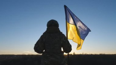 Ukraynalı asker kadın gün batımında mavi-sarı bayrak sallayarak tepenin zirvesinde duruyor. Kamuflaj üniformalı genç bayan asker Ukrayna bayrağını kaldırıyor. Rus istilasına karşı direniş.