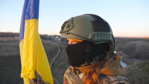 軍事ヘルメットとバラクラバの若い女の子は 手を振って国旗を保持しています ウクライナ軍の女医は田舎で青黄色の旗を持ち上げた 領土のウクライナの概念の侵略 閉じろ — ストック動画