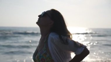 Güneş gözlüklü tatlı esmer, okyanus kıyısında yürüyüp tatil köyünde eğleniyor. Yaz tatili boyunca kıyıda gezinen çekici bir kadın profili. Seyahat ya da tatil kavramı. Yavaşla..