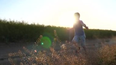 Sahada koşan mutlu bir çocuk ve gün batımında sevinen bir yaz. Küçük çocuk tarlada koşuyor ve toprak yolda toz izi bırakıyor. Doğayla eğlenen bir çocuk ve parlak bir güneş. Yavaşla