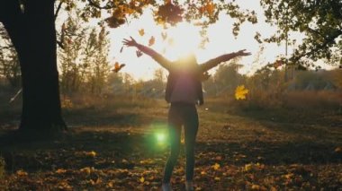 Genç bir kadın ellerini kaldırıyor ve neşeli sonbahar yaprakları etrafında dönüyor. Mutlu kız arka planda günbatımında neşeli duygular gösteriyor. Bayan sonbahar doğasının tadını çıkarıyor. Yavaş çekim. Yakın çekim.