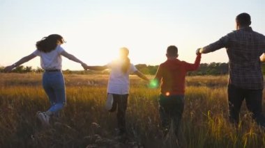 Çocuklu mutlu ebeveynler çim tarlasında el ele tutuşup kollarını kaldırarak koşuyorlar. Anne ve baba ve iki çocuk gün batımında doğanın tadını çıkarıyorlar. Aile mutluluğu kavramı.