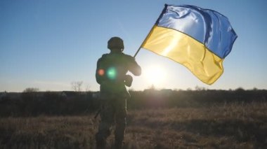 Ukrayna ordusunun erkek askeri ulusal bayrağı kaldırdı. Kamuflaj üniformalı genç adam gün batımında çayırda Ukrayna bayrağı sallayarak koşuyor. İstila direnci konsepti. Arka görünüm.