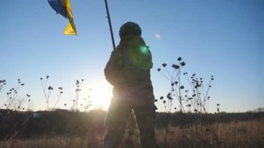 Ukrayna 'nın bayrağını gün batımının arka planında sallayan askeri üniformalı genç bir kız resmi. Ukraynalı kadın asker ulusal bayrağı kaldırıyor. Rus saldırısına karşı zafer