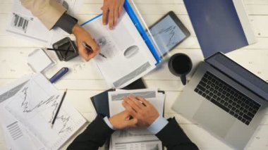 İki başarılı iş ortağının erkek elleri grafikleri inceler ve ofiste finansal raporları tartışırlar. Genç işadamları masada oturuyor ve yeni bir iş projesi geliştiriyor. Üstte görüntü.