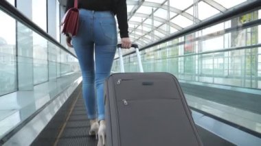 Genç bir kadın bavuluyla terminal koridorundan geçiyor. Yüksek topuklu giyen, yürüyen merdivende yürüyen ve tekerlekli bavulu yuvarlayan tanınmamış bir kız. Bayan seyahate çıkıyor. Arka plan Yavaş çekim Kapat.