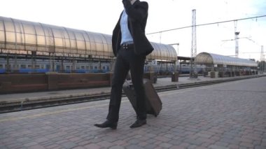 Yakışıklı bir işadamı bavuluyla tren istasyonunda yürüyor ve telefonla konuşuyor. Genç bir iş adamı demiryolu yakınlarında geziniyor ve iş görüşmesi yapıyor. İş gezisi kavramı. Yavaş çekim.