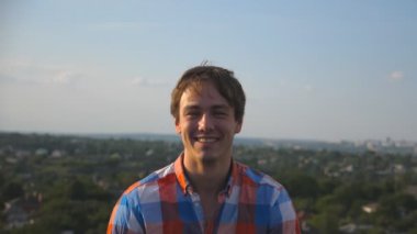 Cityscape arka plan, çatıda oturan genç mutlu adam portresi. Gülüyor ve yüzünde sevinç ifade gösterilen yakışıklı olan. Mutlu bir insan samimi duyguların. Dolly shot ağır çekim yakın çekim.