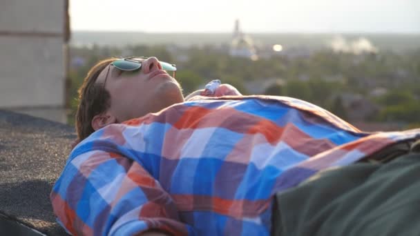 戴着太阳镜的英俊男子躺在屋顶上 背景是城市景观 年轻人在户外放松 享受这一刻 美丽的风景 多利射门慢动作关门 — 图库视频影像