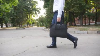 Evrak çantasıyla şehir parkından geçen tanınmamış iş adamlarının bacakları. Başarılı bir şirket çalışanı ofiste yoğun bir iş gününden sonra açık havada yürür. Yavaş çekim kenar görünümü.
