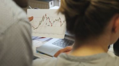 Satış sonuçlarını kağıt üzerinde karşılaştıran genç iş arkadaşları ofisteki bilgisayar ekranında grafikle karşılaştırıyorlar. Profesyonel yöneticiler iş toplantısı sırasında mali raporları inceliyor. Ön planda bulanıklık. Arka görünüm.