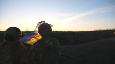 Ukrayna ordusunun askerleri günbatımının arka planına karşı Ukrayna bayrağı çekiyorlar. Askeri üniformalı insanlar sarı-mavi bayrak kaldırdı. Rus saldırısına karşı zafer. İstila direnişi kavramı.