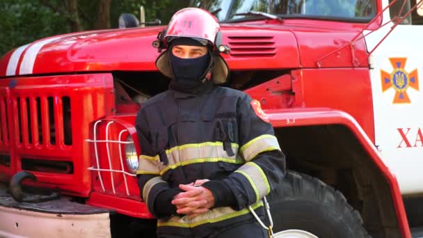 身穿头盔和制服的男性消防员站在一辆红色大轿车旁边的画像 年轻而自信的消防员 装备齐全 在消防车的背景下摆姿势 拯救生命的概念 — 图库视频影像