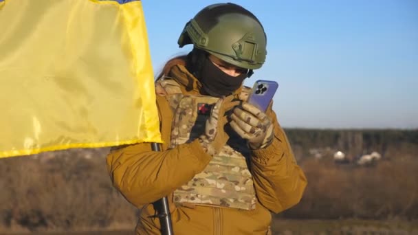 女性のウクライナ軍兵士は 家族とのニュースやメッセージを読むためにスマートフォンを使用しています 軍の制服とヘルメットの女の子は ウクライナの旗を振って保持 ロシアの侵略に対する勝利 閉じろ — ストック動画