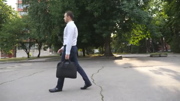 都市の通りに行く正式な摩耗で野心的なハンサムなビジネスマン カメラは成功した若い男を追跡するブリーフケースを持って街の外を歩く オフィスの仕事の概念 サイドビュースローモーション — ストック動画