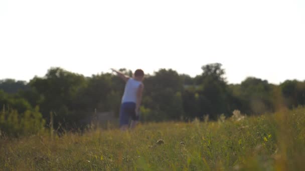 晴れた日には畑の緑の芝生の上を手を上げて育った少年 芝生の屋外で子供のジョギング 夏の牧草地で自然を楽しんでいる男性の子供 スローモーションリアビューを閉じる — ストック動画