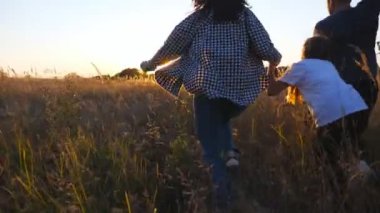 Anne ve baba ve küçük kızı gün batımında çimen tarlasında çok renkli bir uçurtmayla koşuyorlar. Küçük tatlı bir kızı olan genç ebeveynler yaz otlaklarında koşuştururken el ele tutuşuyorlar. Arka görünüm