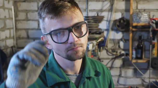 防護眼鏡を外し 額から汗を拭く作業服のプロの整備士 ガレージでカメラを見て疲れた若い修理工の肖像画 メンテナンスサービスコンセプト — ストック動画