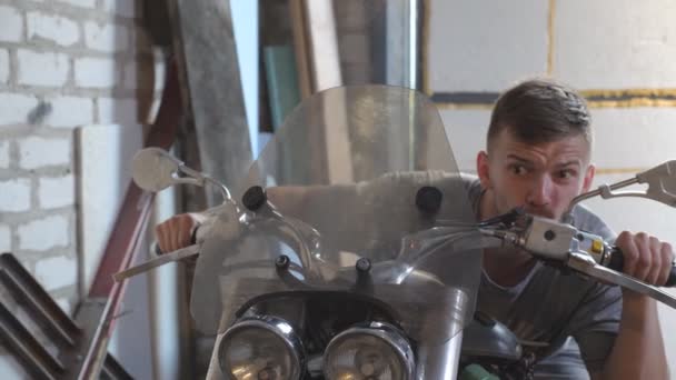 年轻男子假装在车间里骑了一辆摩托车 快乐的家伙在小木屋里骑摩托车鬼混 快乐的人类享受和在车库的乐趣 特写慢动作 — 图库视频影像