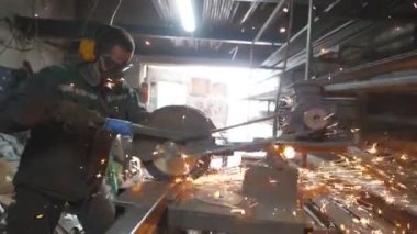 Garajda çeliği kesen dairesel testereli bir adam. Güvenlik koruması altında çalışan işçiler metal kesmek için testere kullanıyor. Metal öğüten adam. Endüstriyel üretim. Sıcak metalden kıvılcımlar çıkıyor. Dolly vurdu..