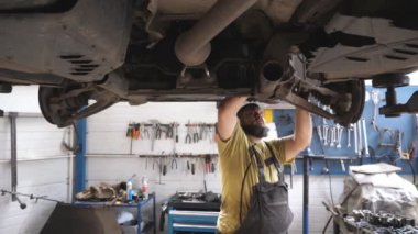 Üniformalı bir tamirci atölyedeki ingiliz anahtarı aletini kullanarak araba tamir ediyor. Garajda kaldırma aracının altında çalışan profesyonel sakallı oto tamircisi. Otomobil bakımı kavramı. Yavaş çekim.