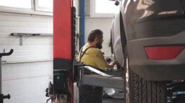 Üniformalı bir tamirci atölyede ya da garajda araba arızalarını tamir ediyor. Araba servisinde kaldırma aracıyla çalışan profesyonel bir tamirci. Düzenli otomobil bakımı kavramı. Yavaş çekim.