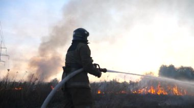 Üniformalı erkek itfaiyeci, kırsal bölgelerde yangın hortumuyla yanan kuru otları söndürüyor. Tam teçhizatlı profesyonel itfaiyeci sahada alev saçıyor. Kahramanlık mesleği ve yangın güvenliği kavramı.