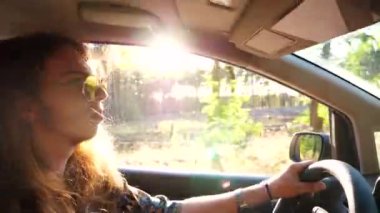 Güneş gözlüklü hippi profiline göre arka planda güneş ışığı olan bir araba kullanıyor. Uzun saçlı, genç hippi bir erkek otomobille kırsala gidiyor. Yolculuk ve seyahat kavramı. Yavaşça kapat..