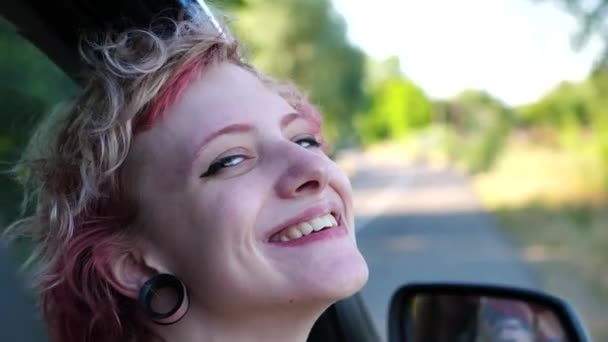 接近一个快乐的朋克女孩的头 她的耳朵从车窗探出 凝视着镜头 微笑嬉皮士女人享受夏天的汽车旅行 冒险和旅行的概念 慢动作 — 图库视频影像