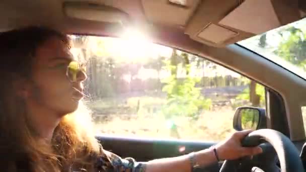サングラスのヒッピーマンが バックグラウンドで日光を浴びた自動車を運転するプロフィール 田舎の道路に自動車に乗って長髪の若い男性ヒップスター 旅と旅のコンセプト スローモーションを閉じる — ストック動画