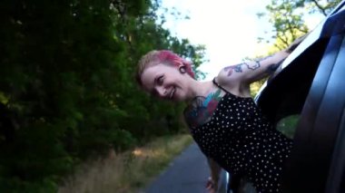 Araba camından sarkan dövmeli mutlu punk kızı ve kır yolunda gezintiden zevk alıyor. Pembe saçlı neşeli hippi kadın araba yolculuğunda eğleniyor. Seyahat konsepti. Yavaş çekim.