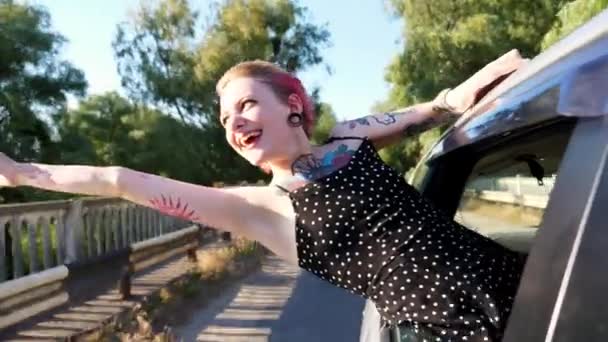 快乐的朋克女孩 身上有纹身 从车窗里探出头来 在路上玩得很开心 快乐的嬉皮士女人 一头粉红的头发 一边开车穿过乡间小路一边享受旅程 旅行概念 — 图库视频影像