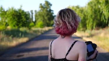 Pembe saçlı mutlu bayan hippi boş kır yolundaki fotoğraf makinesinde fotoğraf çekiyor. Kırsal evin önünde kameraya poz veren dövmeli, kaygısız bir punk kızı. Hippi kadın dışarıda resim çekiyor..