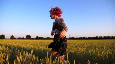 Gün batımında buğday çayırında koşan dövmeli mutlu punk kadın. Yeşil arpa tarlasında koşarken pembe saçlı neşeli hippi kız eğleniyor. Neşeli bayan hippi Yazın tadını çıkarıyor.