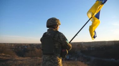 Soldier of ukrainian army stands at hill and holds waving flag of Ukraine. Man in military uniform and helmet lifted up flag against sunset. Victory against russian aggression. Invasion resistance.