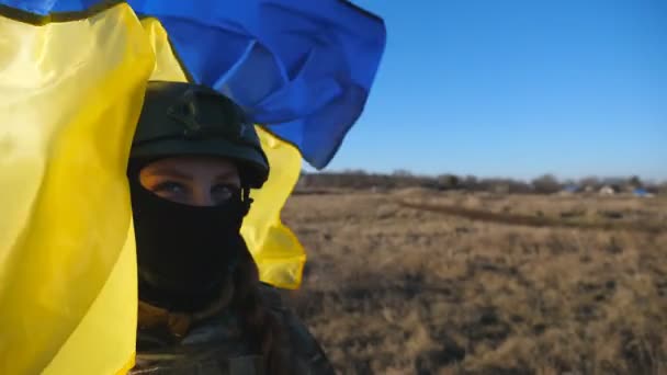 ウクライナ軍の女性軍医は 青黄色のバナーを振ってカメラを見ています 晴れた日にフィールドでウクライナのフラグを保持カモフラージュ制服やヘルメットで女の子の肖像画 ドリーショット — ストック動画