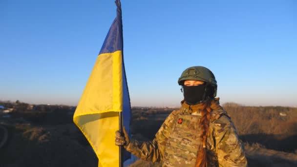 カモフラージュのウクライナ軍の女性は 日没時に青色のバナーを持ち上げています 若い女性兵士はウクライナの旗を振って丘の頂上に立っている 戦争における侵略抵抗と勝利 — ストック動画