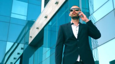 İş adamı güneş gözlüklü, ofisin yakınında yürüyor ve telefonda konuşuyor. Genç mutlu iş adamı dışarı çıkıyor ve cep telefonuyla konuşuyor. İş konuşmasının portresi. Yavaş çekim. Yakın çekim..