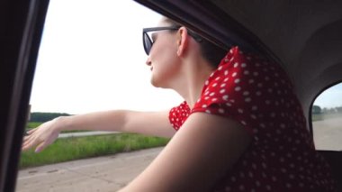 Happy girl in sunglasses leaning out of retro car window and enjoying trip. Attractive woman looking out of moving vintage auto on summer day. Travel and freedom concept. Slow motion Close up.