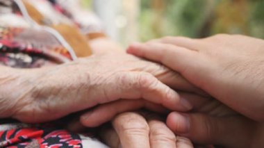 Yaşlı büyükannesinin yaşlı ellerini okşayan genç bir erkeğin yan görüntüsü. Aile dışarıda vakit geçiriyor. Şefkatli ve sevgi dolu bir kavram. Yavaşça kapat..