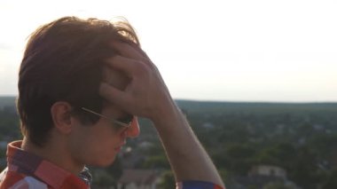 Güneş gözlüklü yakışıklı adam çatıdan manzaraya bakıyor ve saçını düzleştiriyor. Genç adam şehrin manzarasını çatıdan izliyor ve güzel manzaranın tadını çıkarıyor. Dolly vurdu. Ağır çekim..