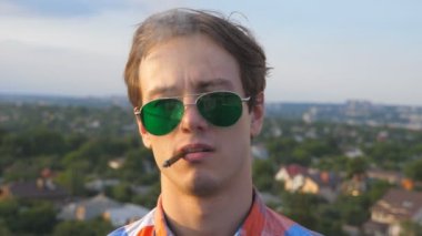 Güneş gözlüklü yakışıklı bir adamın portresi. Çatının kenarında durmuş sigara içiyor. Genç ciddi adam çatıda dinleniyor ve arka planda şehir manzarasıyla saçını düzleştiriyor..