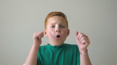Sevimli bebek başarısını kutluyor ve baş parmağını kaldırıyor. Yüzünde neşe ifadesi olan erkek çocuğun duygularını kapat. Kapalı alanda çilleri olan kızıl saçlı yakışıklı çocuğun portresi. Yavaş çekim.