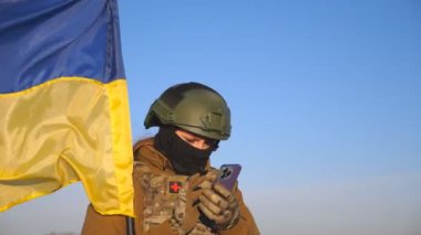 Female ukrainian army soldier using smartphone to read news or messaging with family. Girl in military uniform and helmet holding waving flag of Ukraine. Victory against russian aggression. Close up.