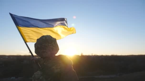 ウクライナの軍服を着た若者が夕日を背景にウクライナの旗を振った ウクライナ陸軍の兵士が国旗を掲げて丘に上がった ロシア侵略の概念に対する勝利 リアビュー — ストック動画