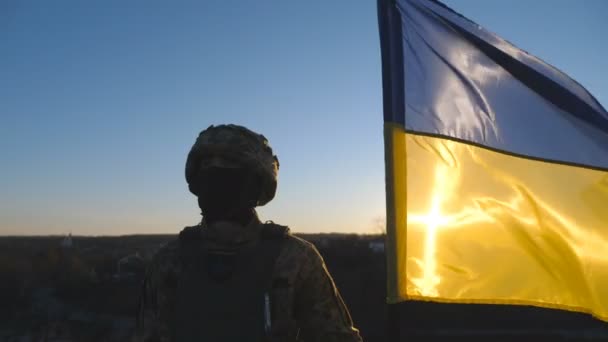 ウクライナ軍のドリー ショットは 夕日を背景に国旗を持ち上げた 軍服とヘルメットの若い男性兵士は 丘の頂上にウクライナの波の旗を保持しています — ストック動画