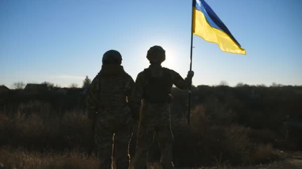 ウクライナの旗が掲げられた丘の頂上に立っているウクライナ軍の女性と男性の兵士 カモフラージュのユニフォームの軍用カップルは 太陽と青い空を見ながらお互いの手を握っています ドリー ショット — ストック動画